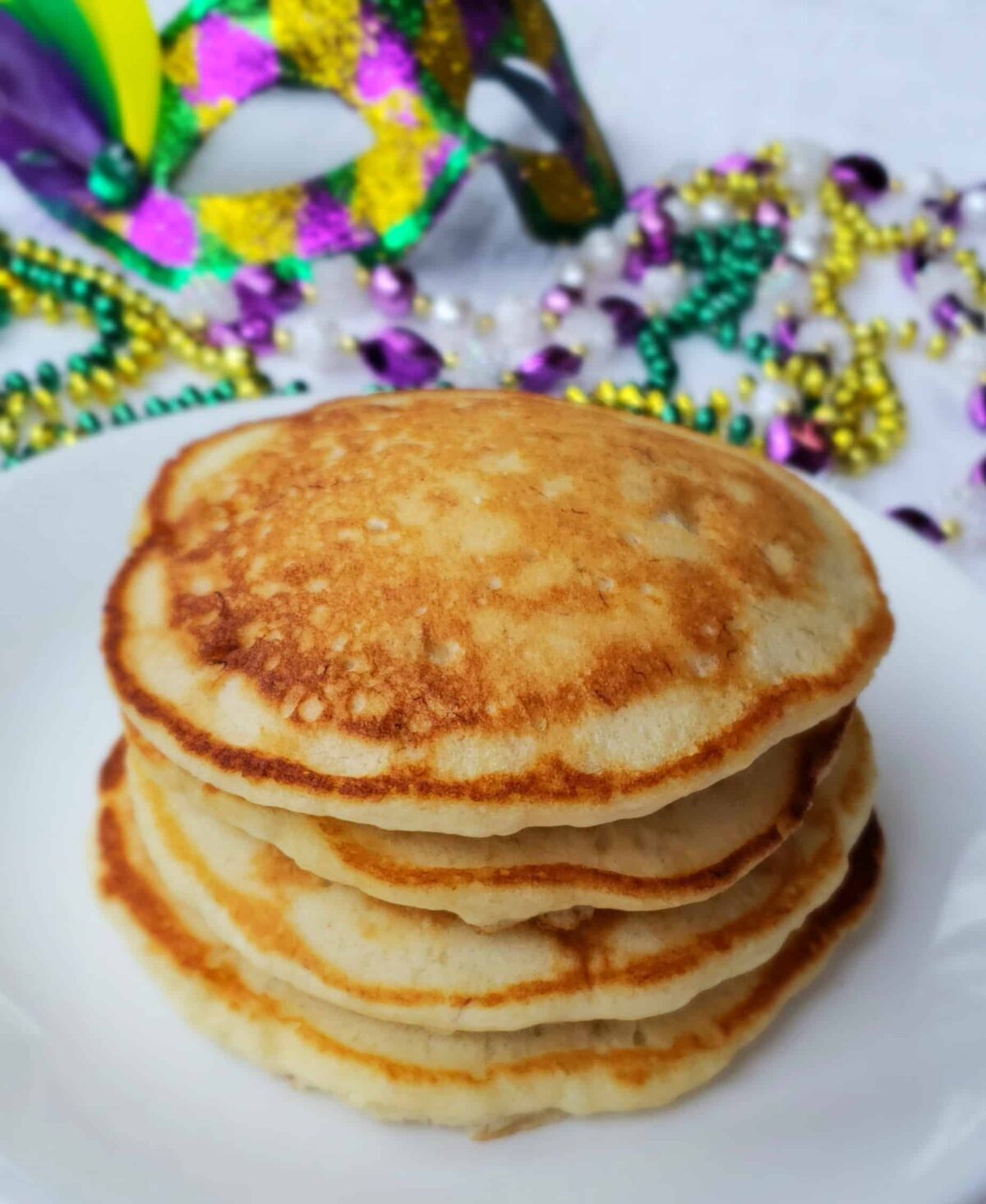  A stack of shortcut banana pancakes before drizzling icing and sprinkles on top. Mardi gras beads in back ground