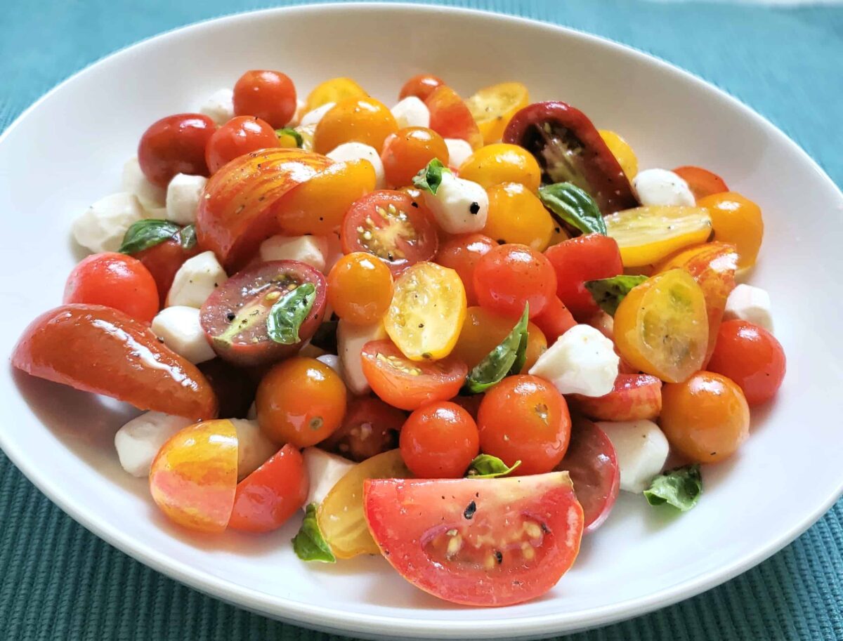  multi colored grape and tear drop tomatoes, pearl mozzarella, and basil in a white bowl