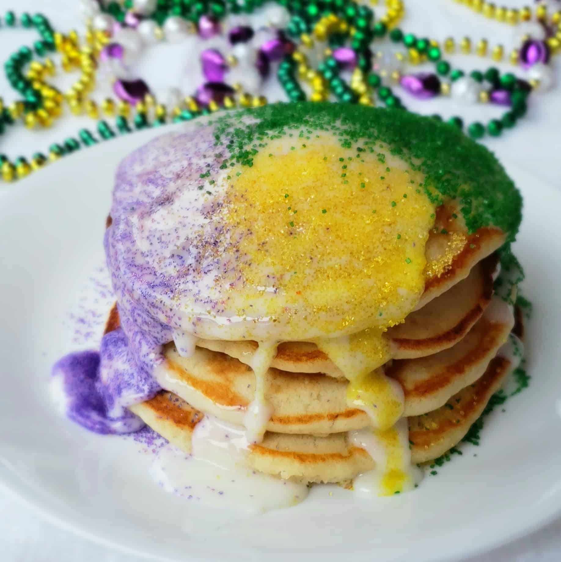 A stack of four pancakes with mardi gras beads in background.Can't make it to Mardi Gras? These shortcut banana pancakes drizzled with icing and sugar sprinkles are perfect for Fat Tuesday- also called Pancake Day!