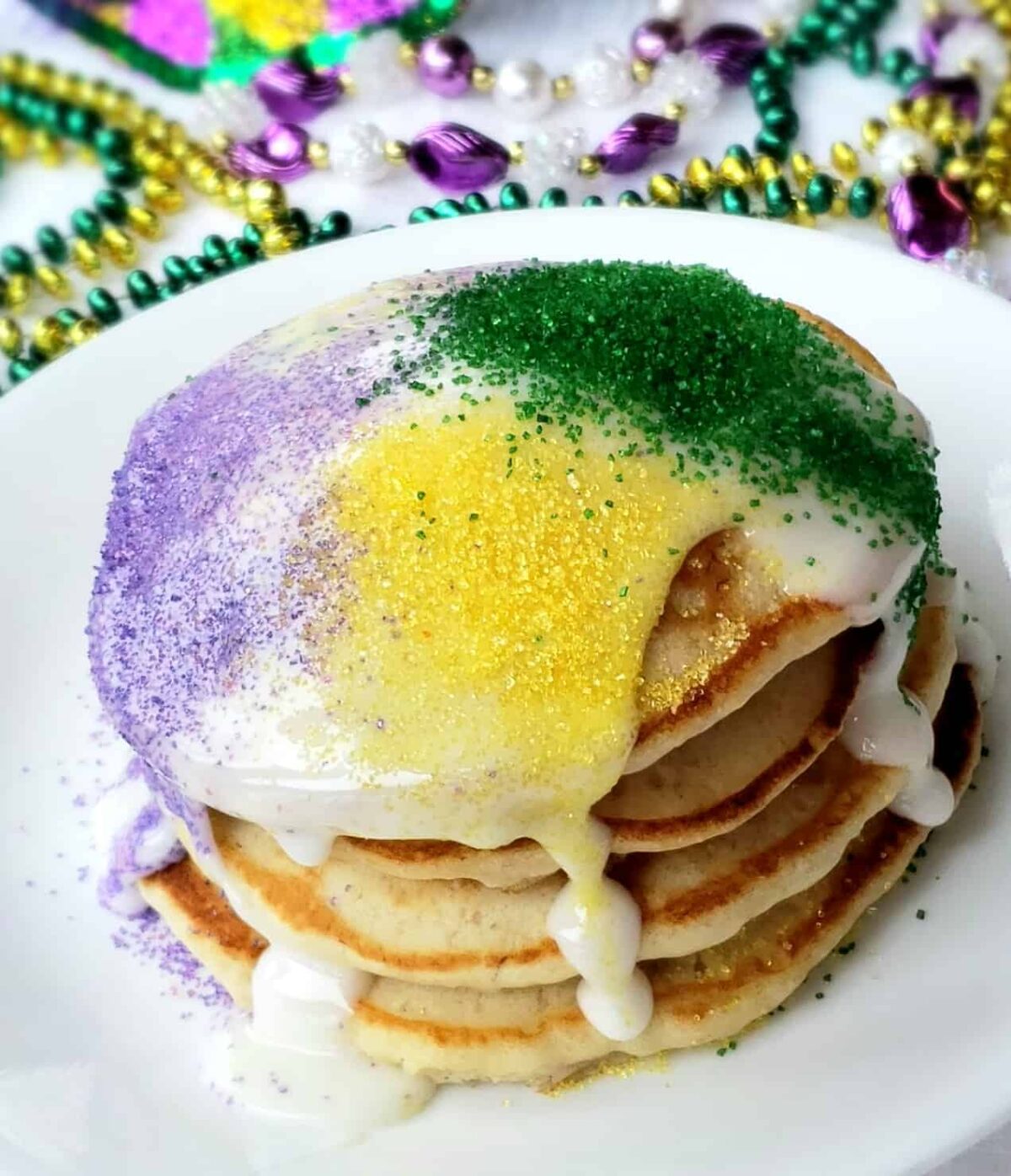 A stack of four pancakes with mardi gras beads in background.Can't make it to Mardi Gras? These shortcut banana pancakes drizzled with icing and sugar sprinkles are perfect for Fat Tuesday- also called Pancake Day!