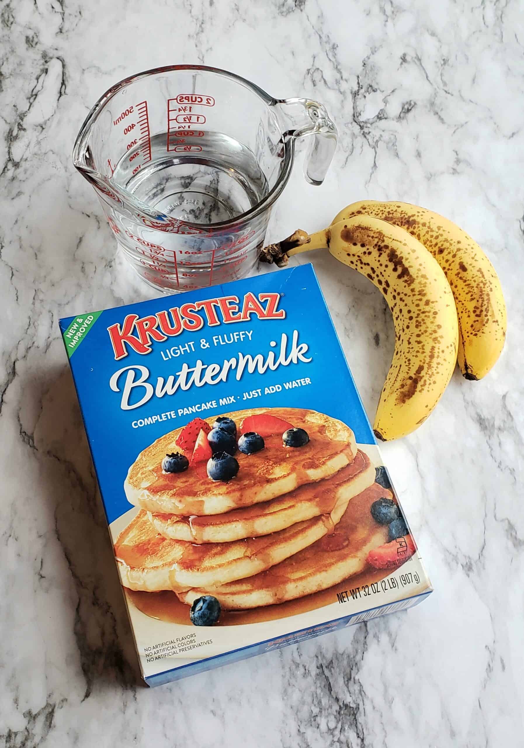  pancake mix Krusteaz and measuring cup of water bananas on marble surface