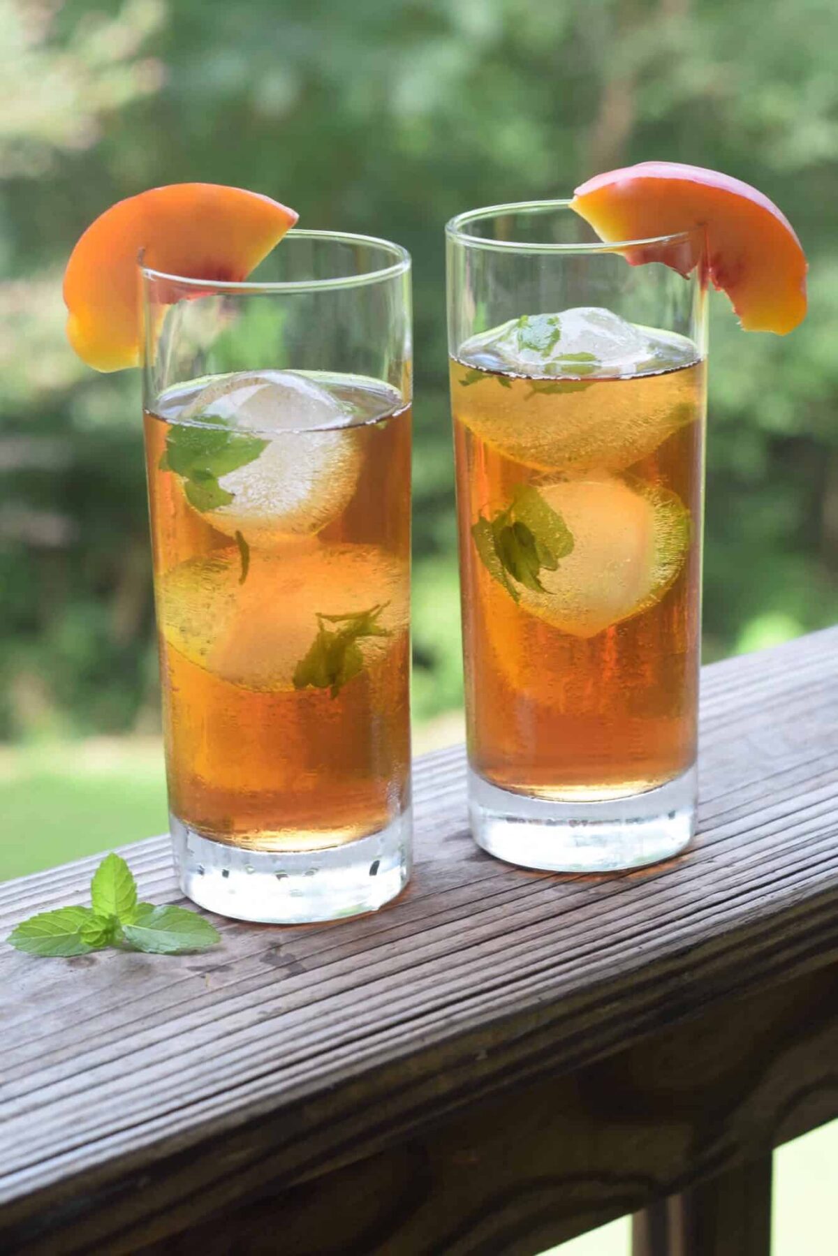 Mint Ice cubes in tall tea glasses with peach wedges on glass. Glasses on wooden railing with greenery in background