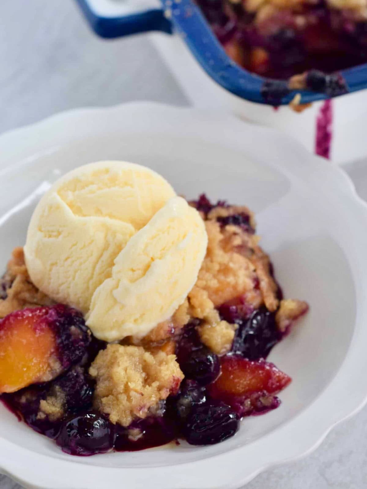 White bowl of peach blueberry cobbler topped with scoop of vanilla ice cream.
