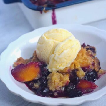 Blueberry Peach Cobbler in a fluted white bowl with scoop of ice cream on top and casserole dish in background.