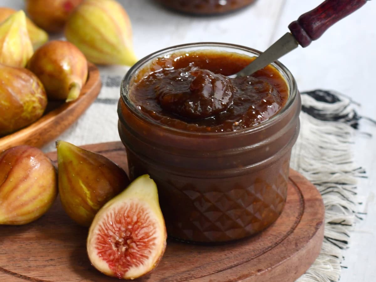 A small open jar of fig jam with a little spoon in it with a fresh fig half leaned on it.