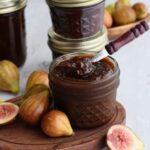 A small open jar of fig jam with a little spoon in it with a fresh fig half leaned on it.