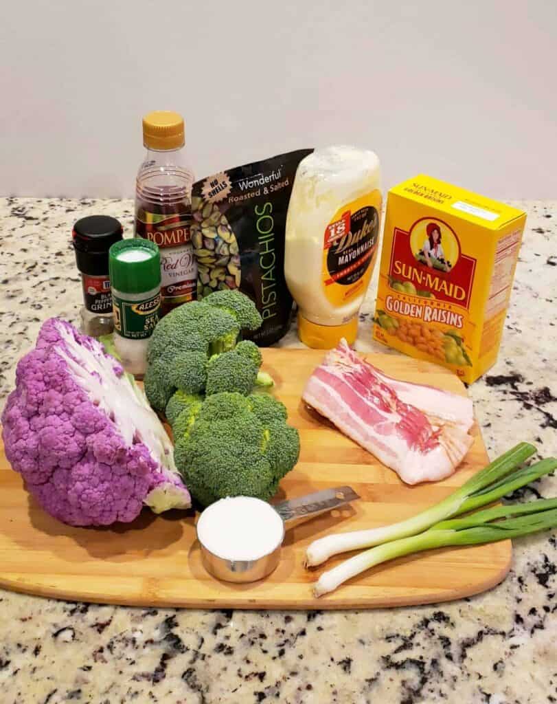 Ingredients on a cutting board: red wine vinegar, Dukes mayonnaise, green onion, golden raisins, bacon, broccoli, purple cauliflower