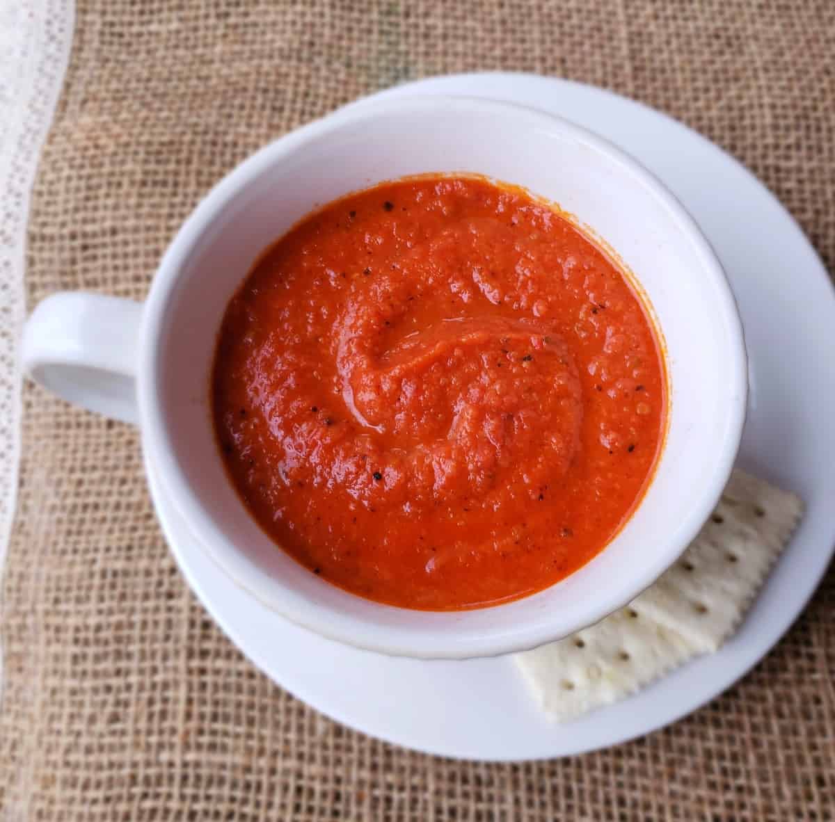 white cup of tomato soup on a white plate with crackers on the side.