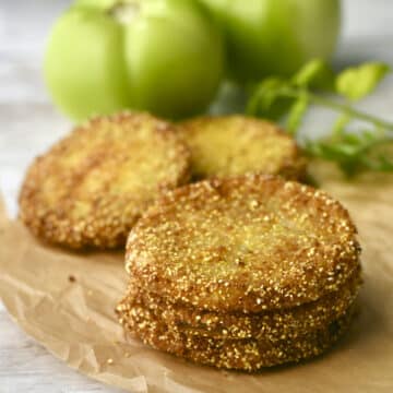 Three thin sliced Southern fried green tomatoes with cornmeal on brown paper with green tomatoes in background.