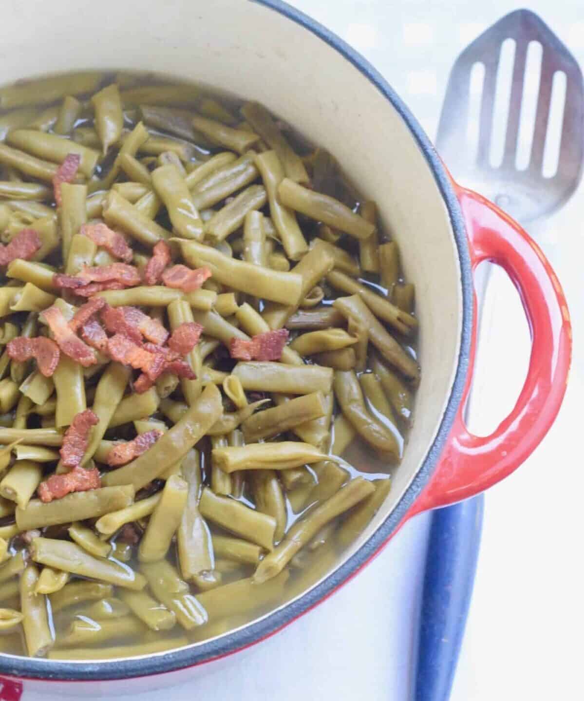 Red pan of green beans with bacon on top with blue slotted spoon on surface.