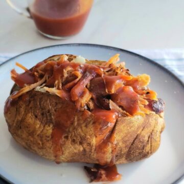 Baked potato stuffed with meat covered in sauce on a white plate. A clear tiny pitcher of bbq sauce in background
