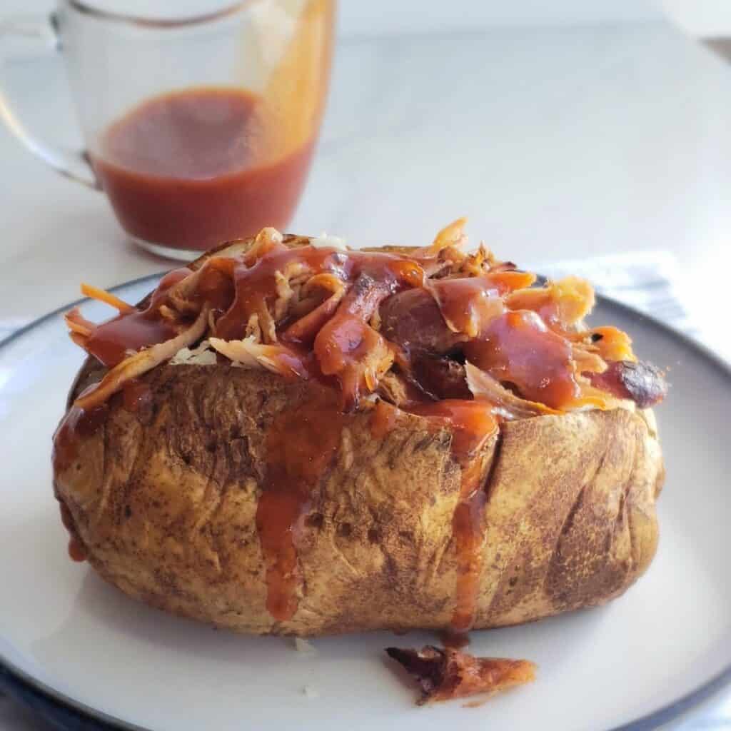 Baked potato stuffed with meat covered in sauce on a white plate. A clear tiny pitcher of bbq sauce in background with sauce dripping down the side