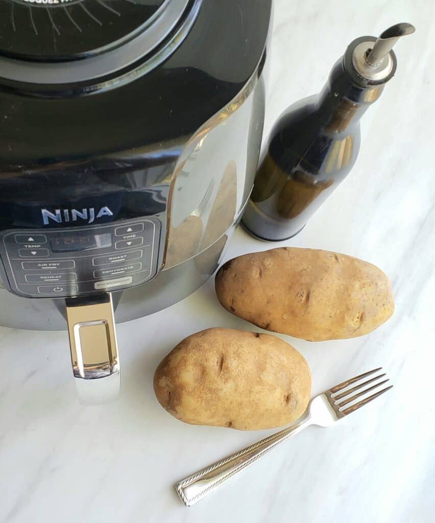 Air fryer, 2 baking potatoes, a fork and an oil dispenser on a white surface