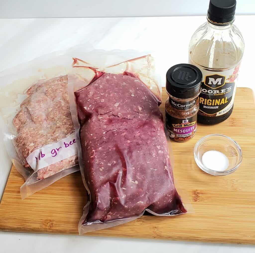 freezer bag of ground beef, one bag of venison, spice bottle Moores seasoning and salt on a wooden cutting board
