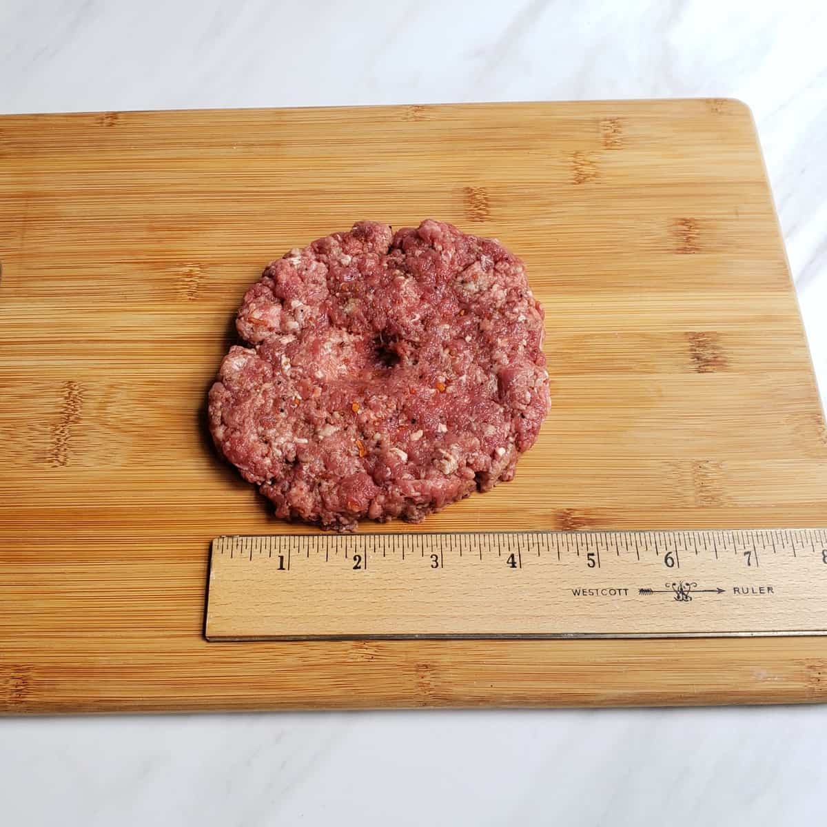 Raw ground meat patty with thumb print pressed in middle and ruler next to it
