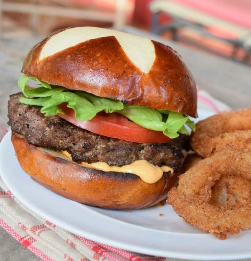 Grilled burger on a pretzel bun with a tomato, green lettuce, pink sauce on a white plate and onion rings peeking in the right side