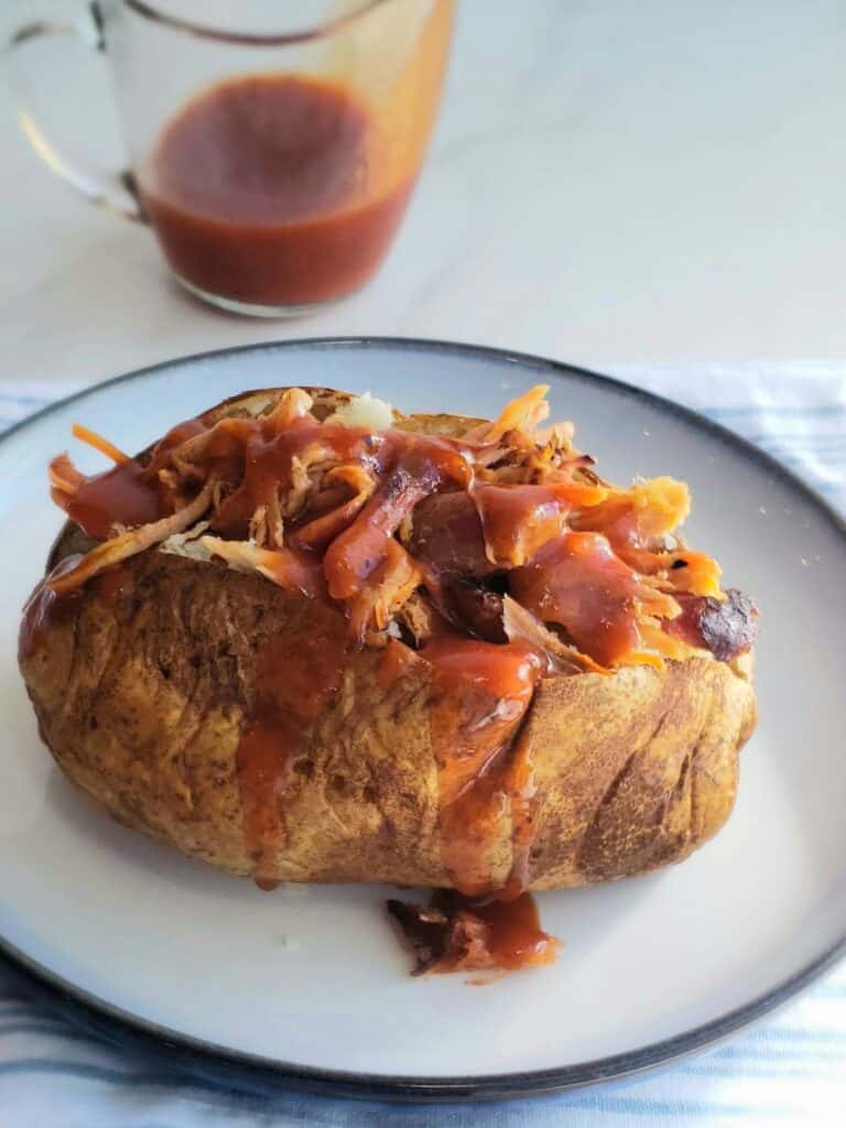 Baked potato stuffed with meat covered in sauce on a white plate. A clear tiny pitcher of bbq sauce in background