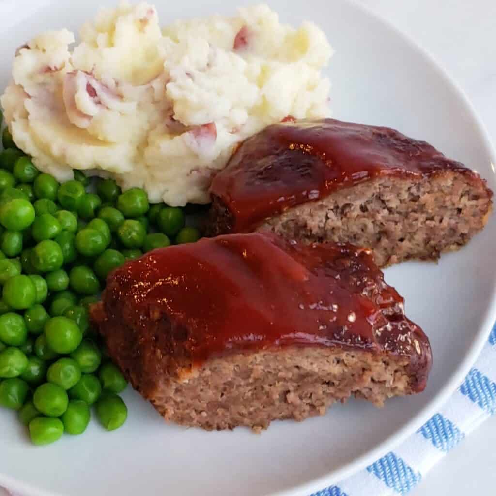 Sliced meatloaf with ketchup sauce on top served on a plate of mashed potatoes and English peas