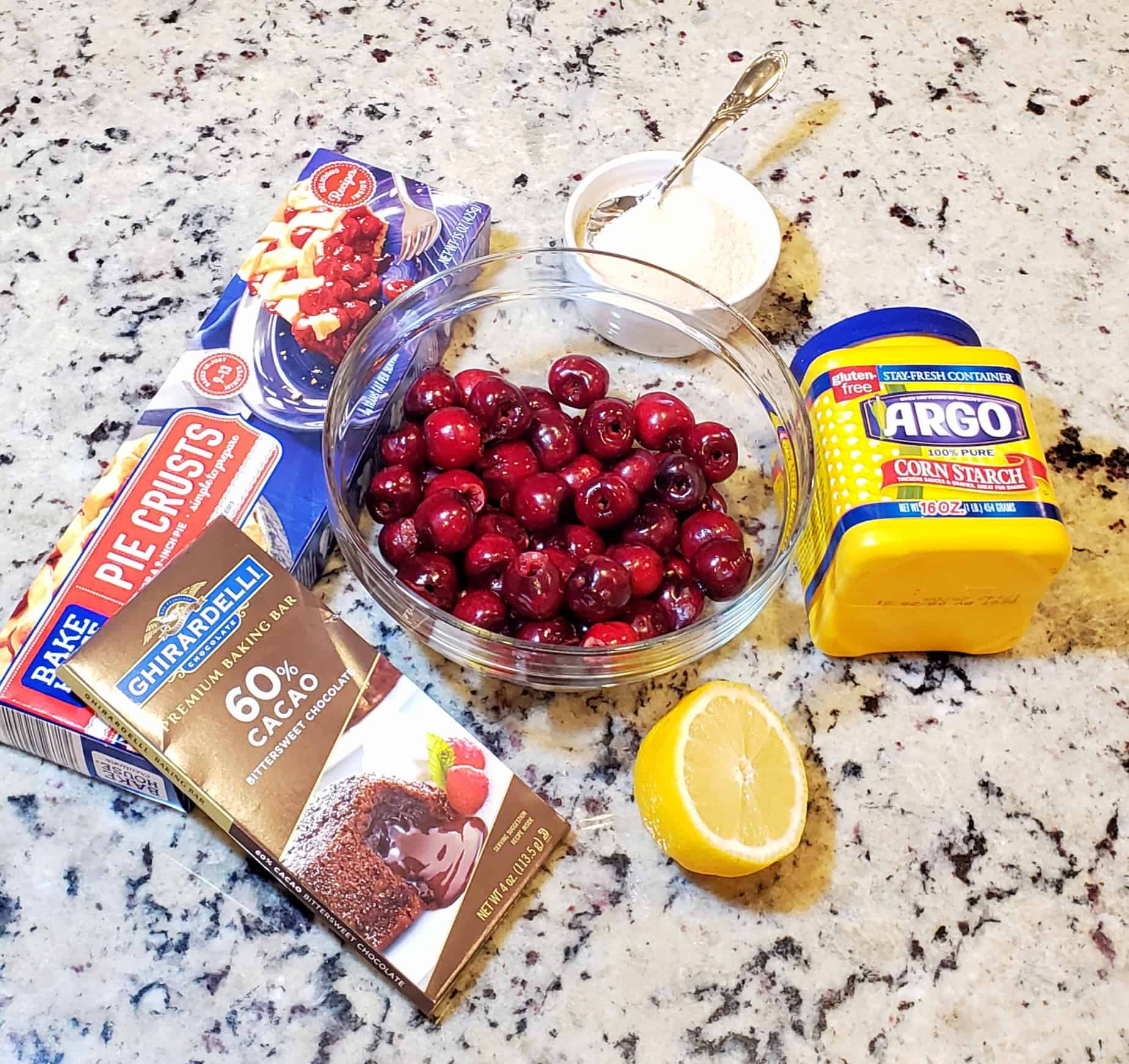 bowl of fresh cherries, lemon, cornstarch, chocolate bar and box of pie crust on a granite surface