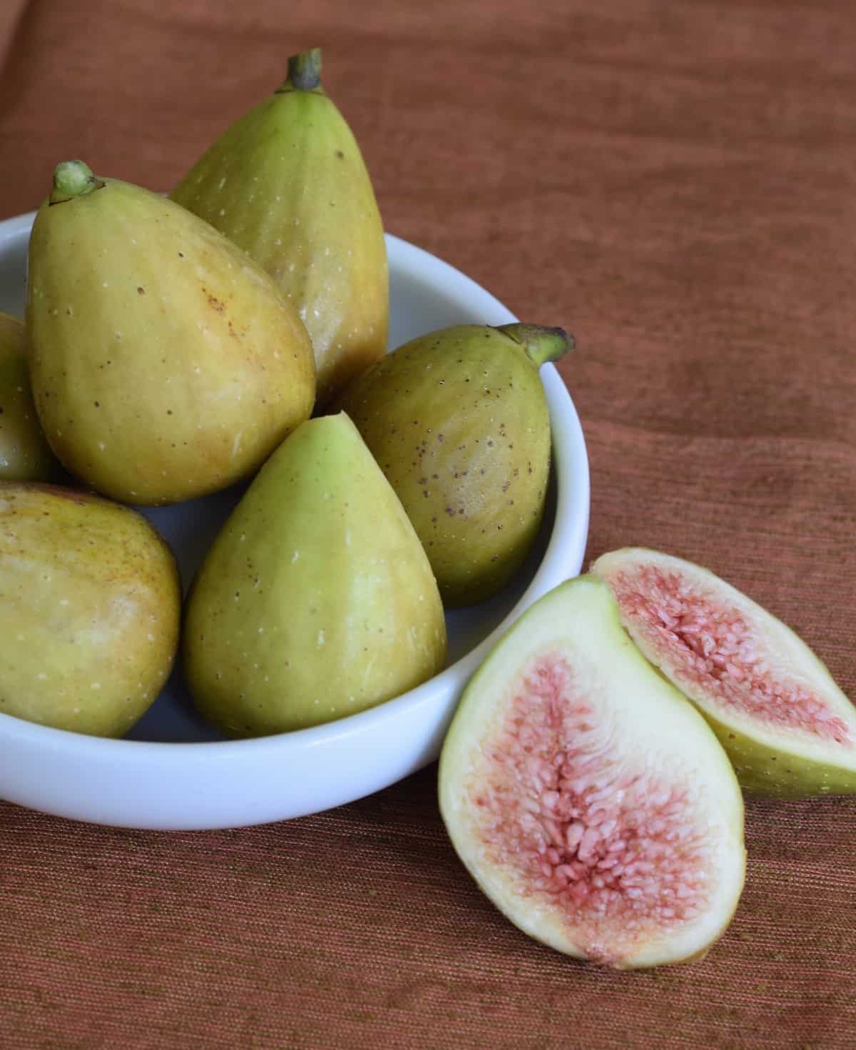 White bowl of green figs with one cut in half on wooden surface.