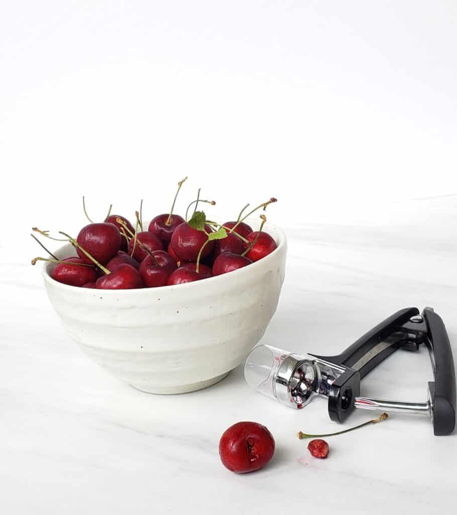 white bowl of bing cherries with stems and cherry pitter