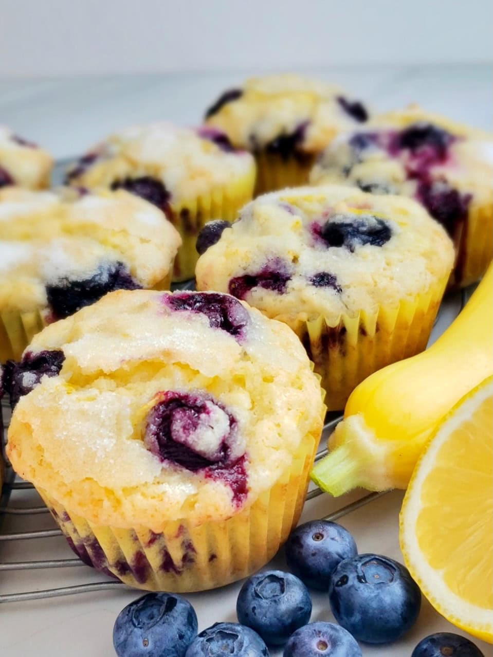 Lemon Blueberry Muffins with paper wrapper open next to a yellow squash and lemon with fresh blueberries.
