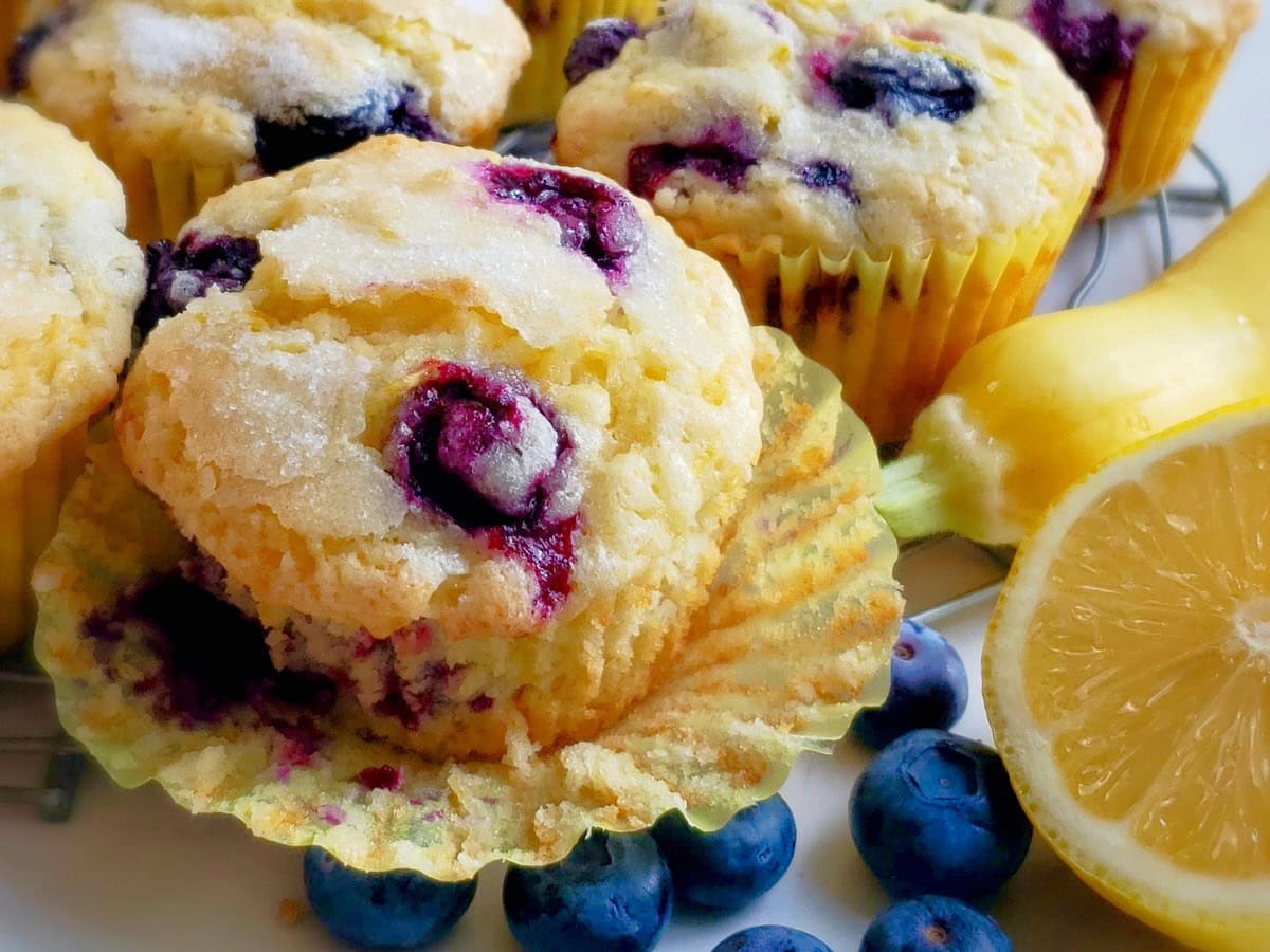 Lemon Blueberry Muffins with paper wrapper open next to a yellow squash and lemon with fresh blueberries.