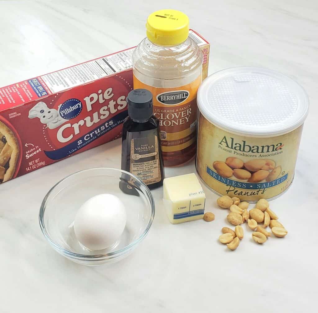 Honey Peanut Pie Tassies Ingredients on marble surface: pie crust dough, honey, egg, peanuts, vanilla butter