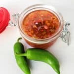 Open hinged jar of red pepper jelly with two jalapenos on white surface