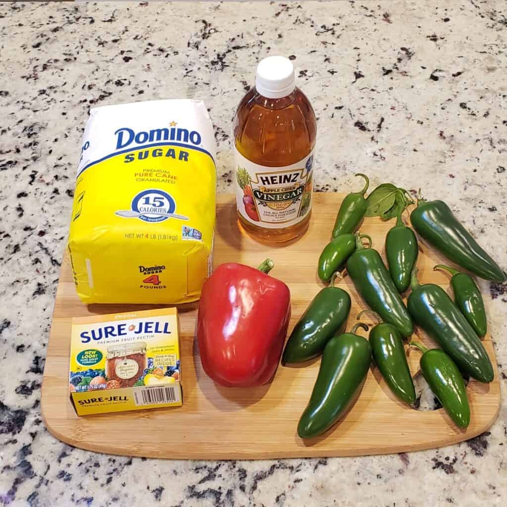 Ingredients for pepper jelly on a wooden cutting board: sugar, Sure-Jell, apple cider vinegar, red pepper, jalapeno peppers