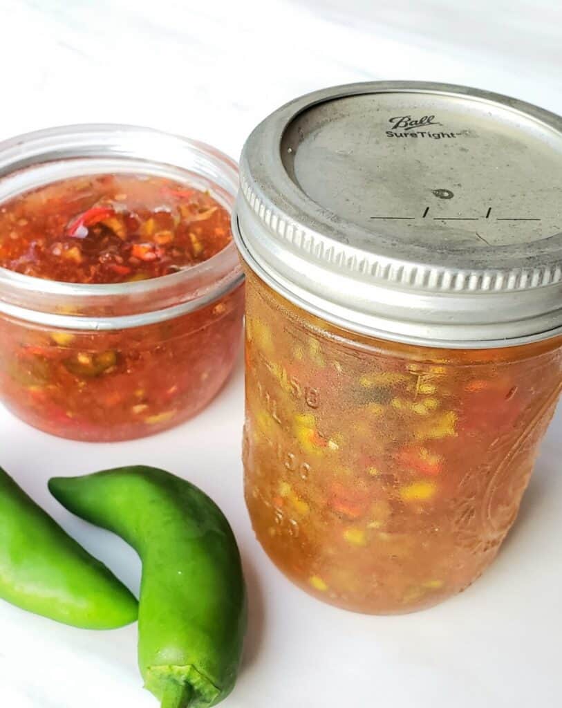 Two jars of pepper jelly; one red and lid off; two jalapenos on surface