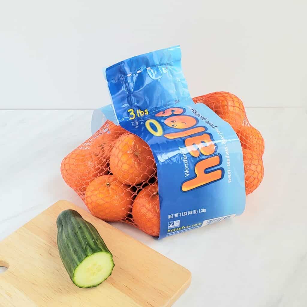 Bag of mandarin oranges and a pied of cucumber on a wooden cutting board