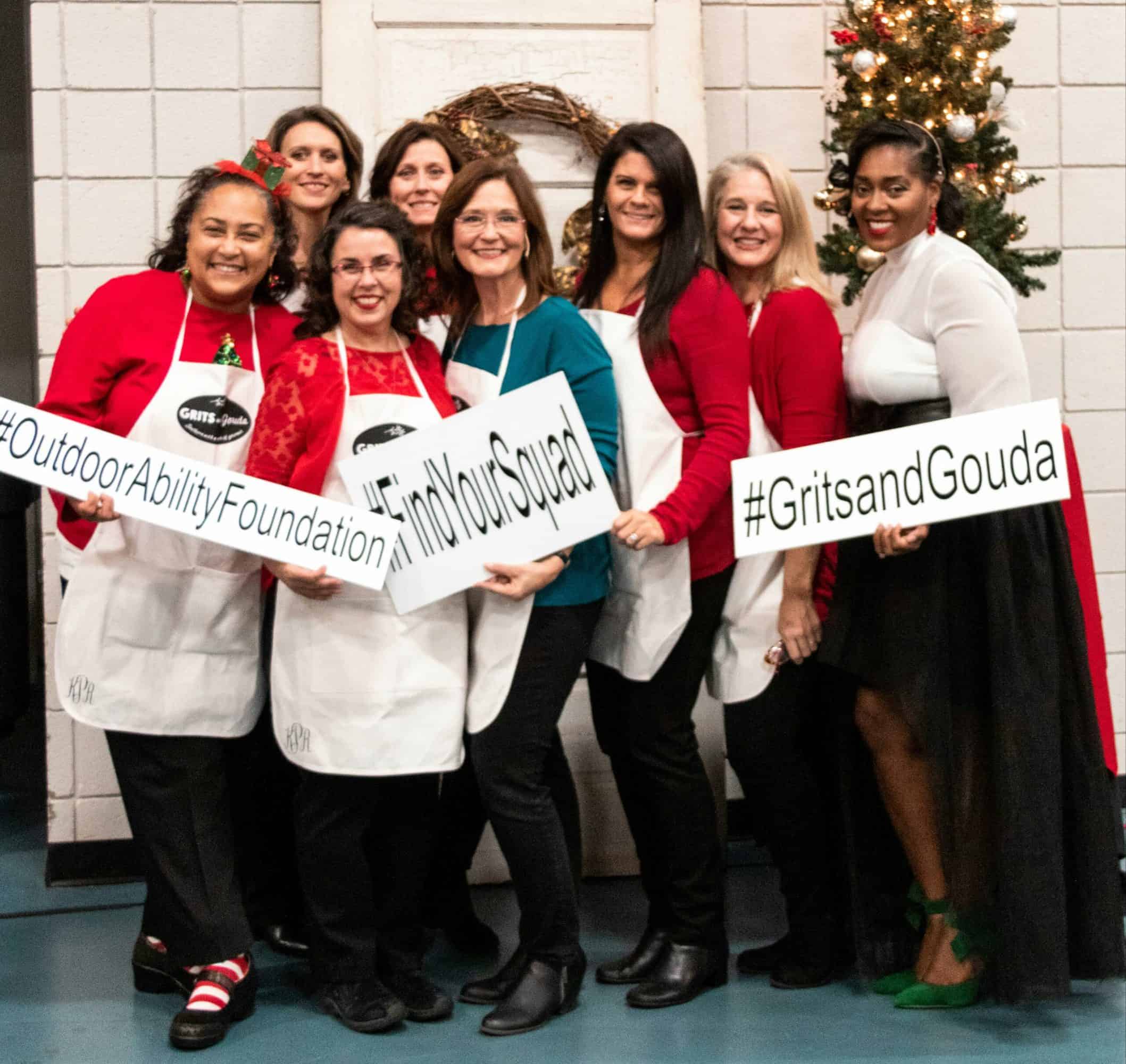 8 women wearing white aprons by Christmas tree holding fundraiser signs