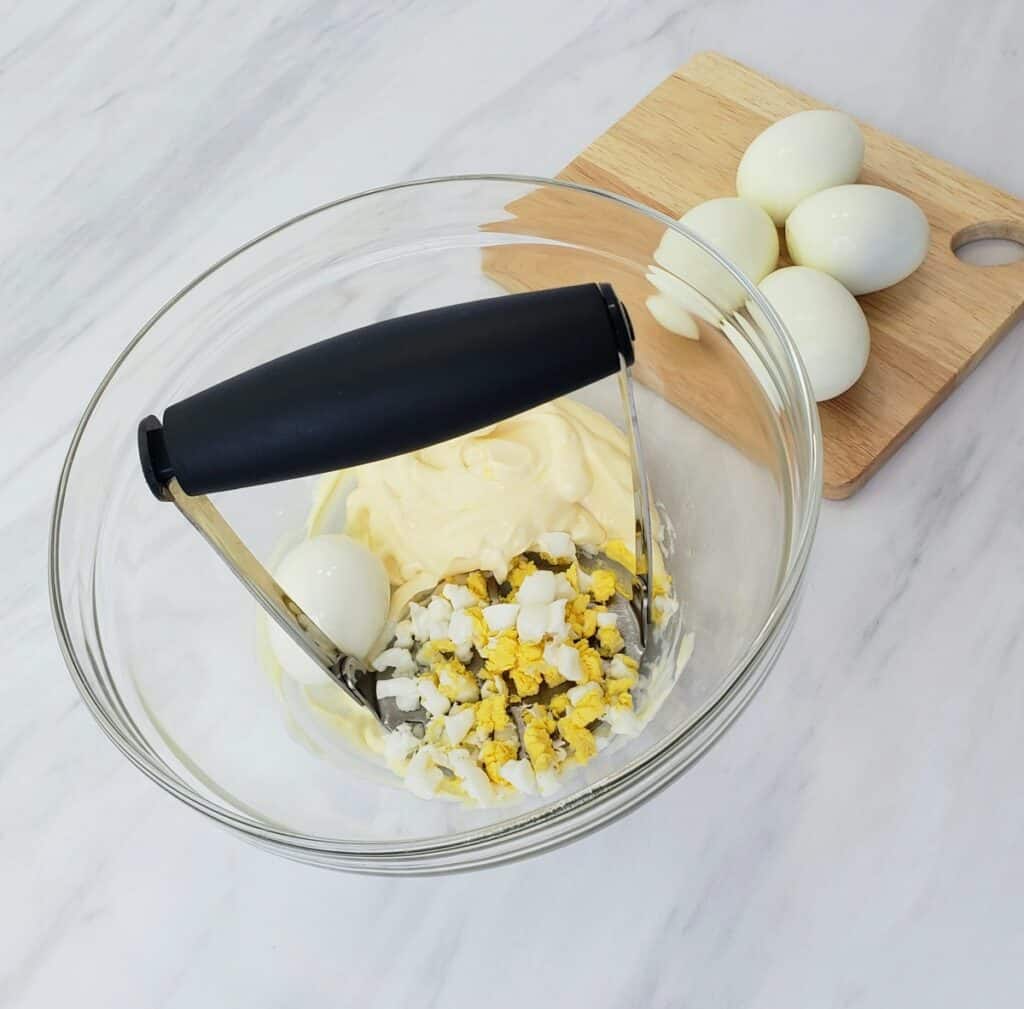 Potato masher mashing boiled eggs in a glass bowl; boiled eggs on a cutting board