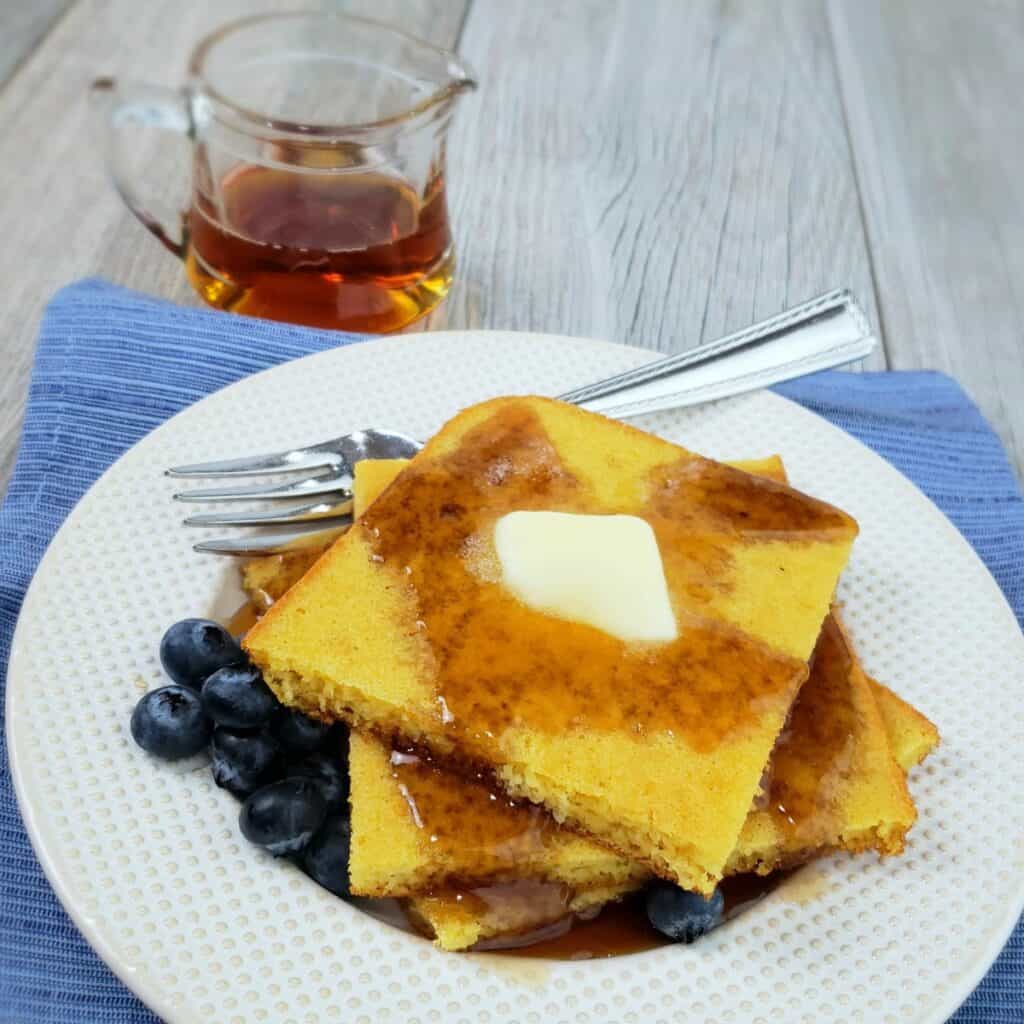 Stack of square pancakes on white plate with butter pat and drizzled syrup; tiny pitcher of syrup in background