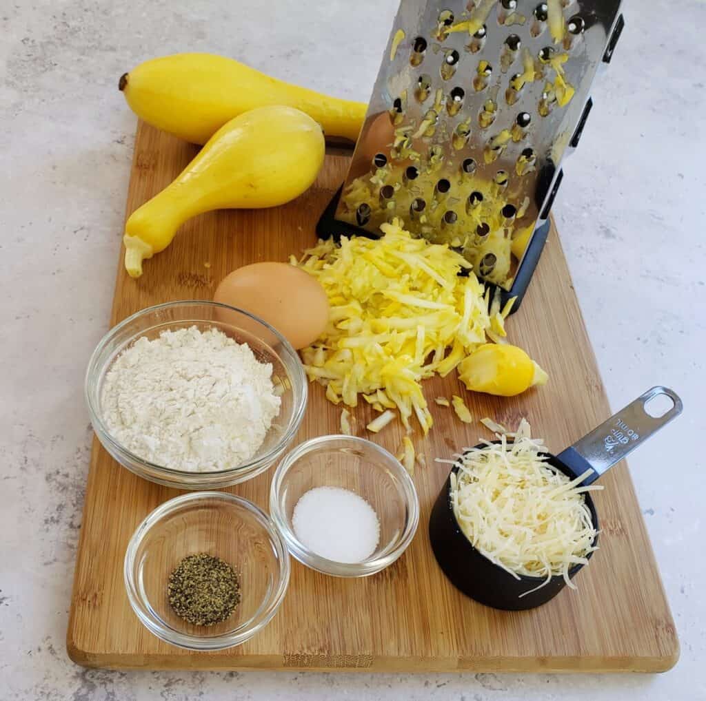 Ingredients on a wooden cutting board: shredded yellow squash, measuring cup of Parmesan cheese, salt, pepper, flour, egg