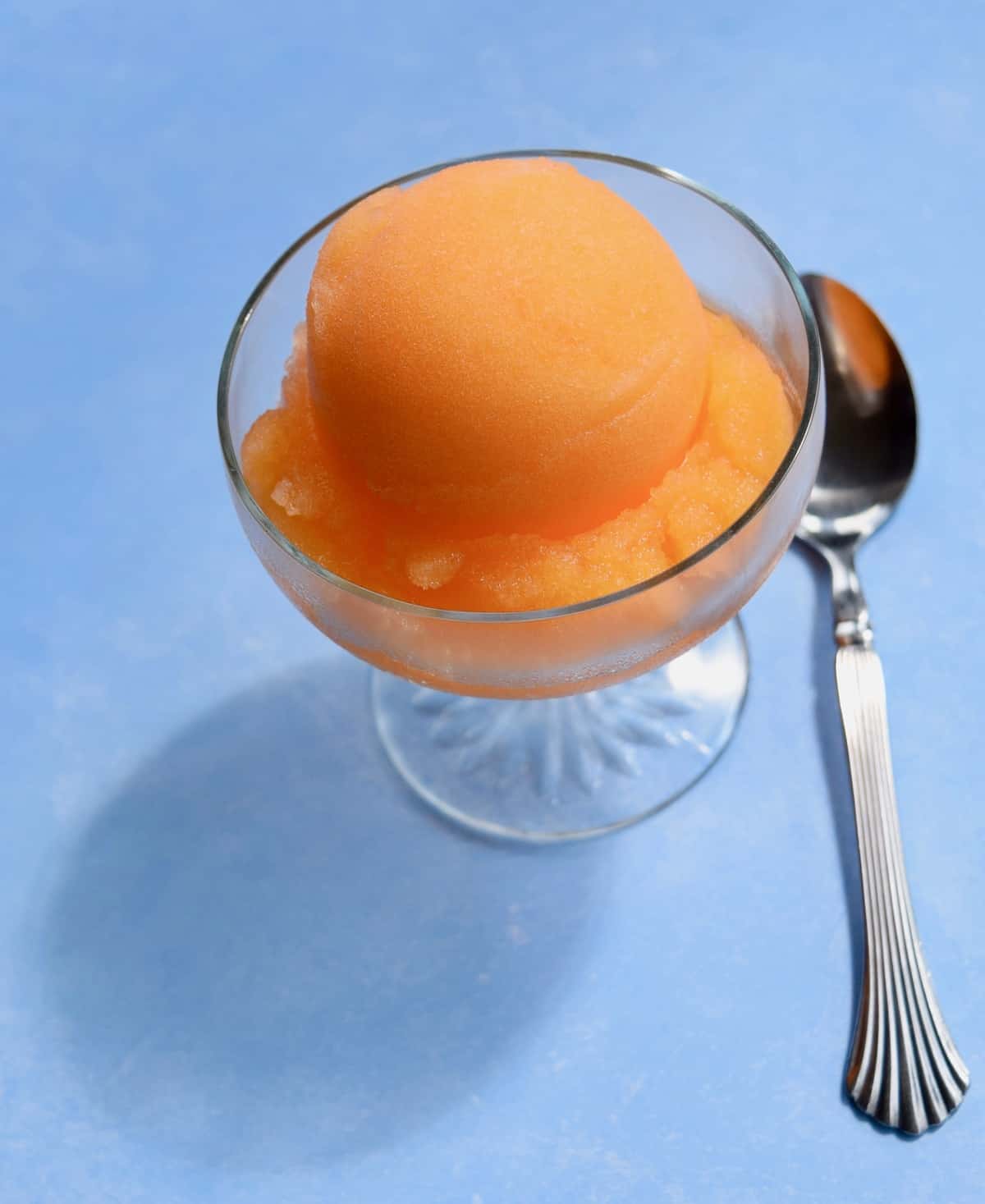 Scoop of orange sorbet casting shadow on blue surface with spoon on right