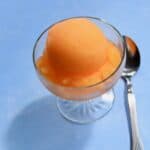 scoop of orange sorbet in a glass dish on blue surface with spoon on surface.