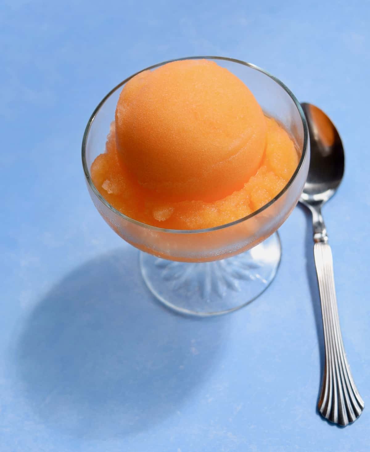 scoop of orange sorbet in a glass ice cream dish on blue surface with spoon on surface.