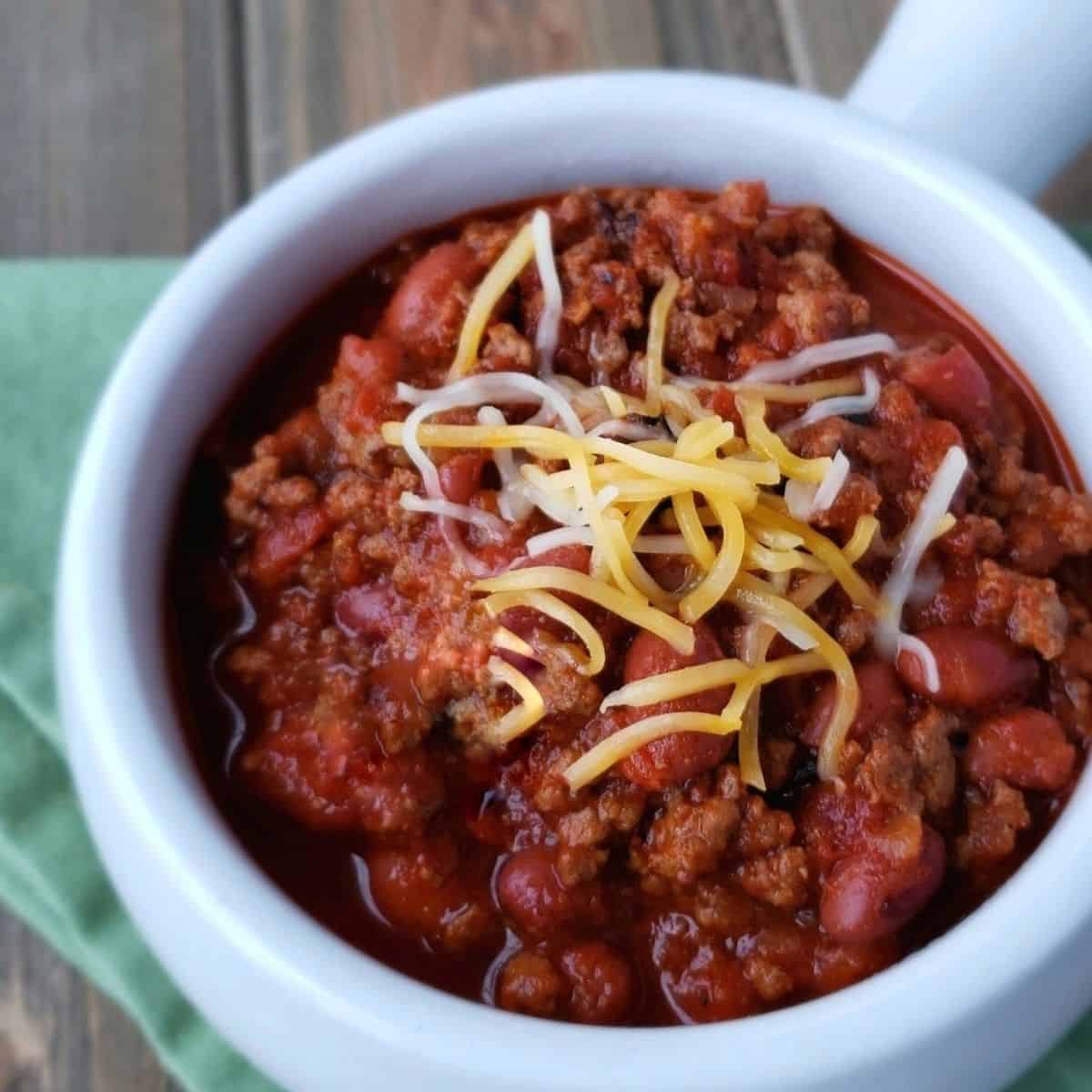 Chili in a white bowl with a handle on a green cloth on a wooden surface