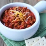 White bow of venison chili topped with cheese on a green napkin with saltine crackers.