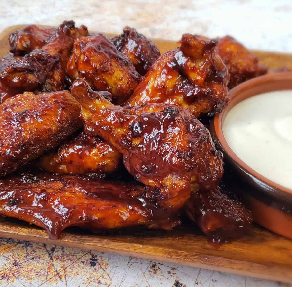 A rectangle wooden platter of bbq sauced chicken wings with bowl of Ranch dressing