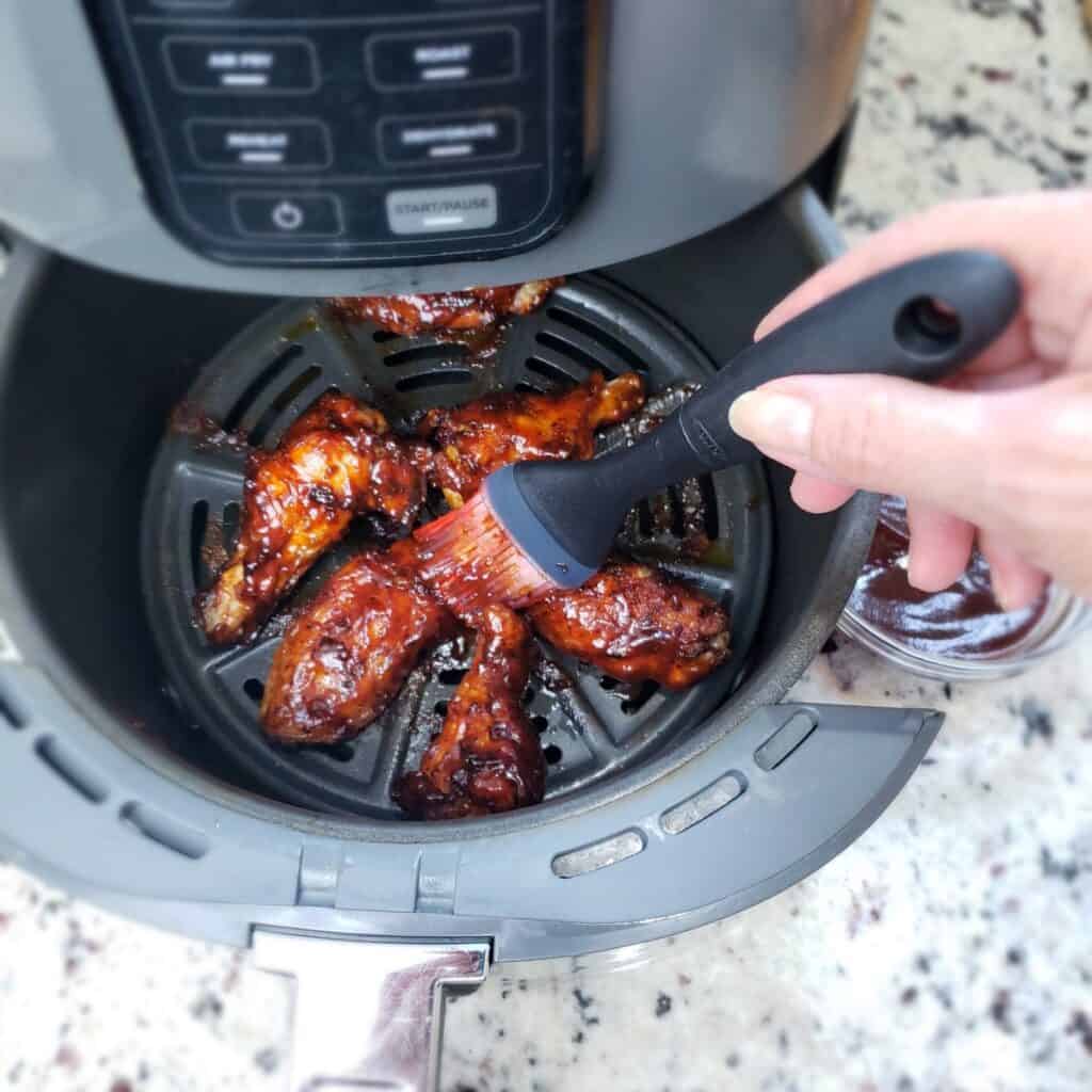 A hand basting bbq sauce on cooked chicken wings with a black pastry brush