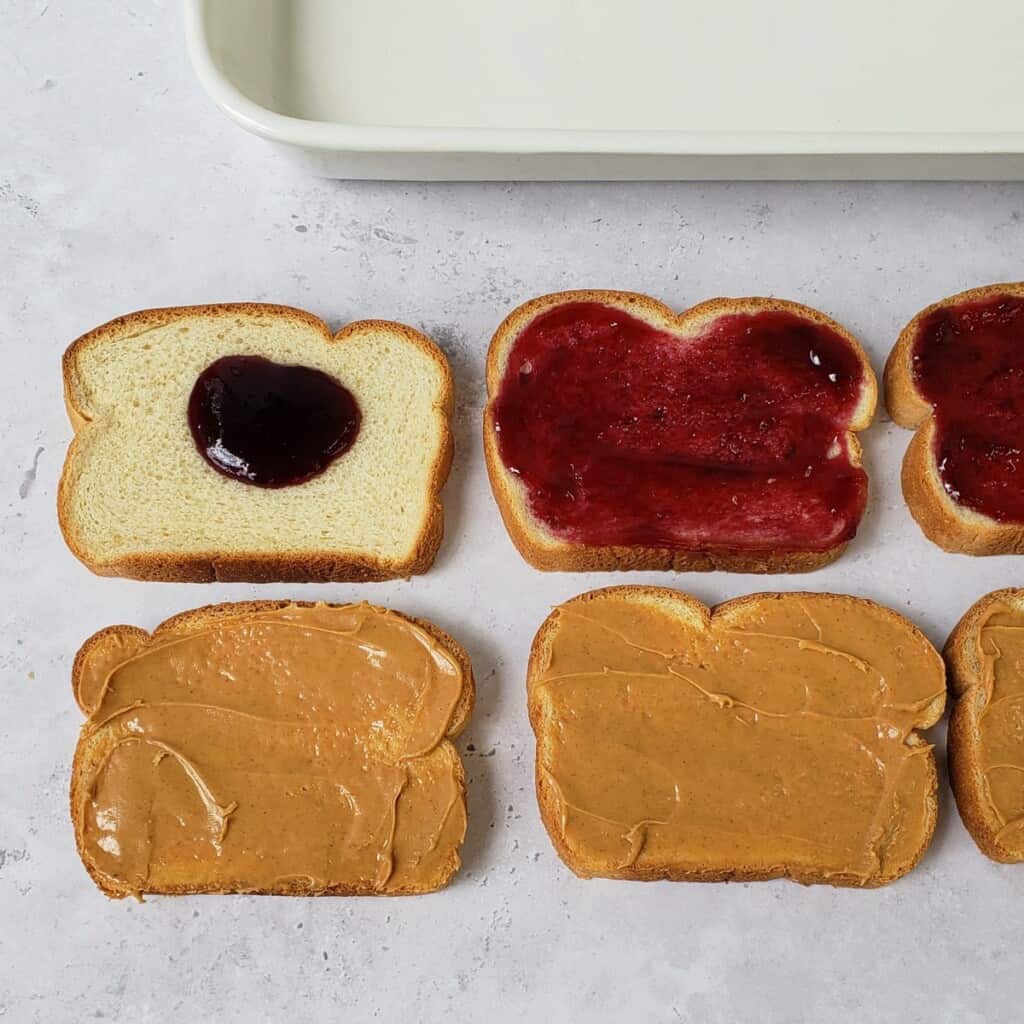 Slices of bread on marble surface with some spread with peanut butter, some with grape jam.