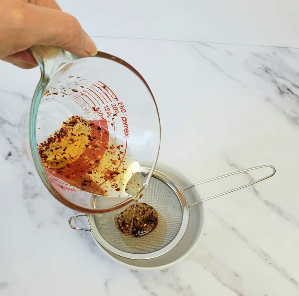 Hand pouring honey and chile pepper flakes from measuring cup into metal sieve.