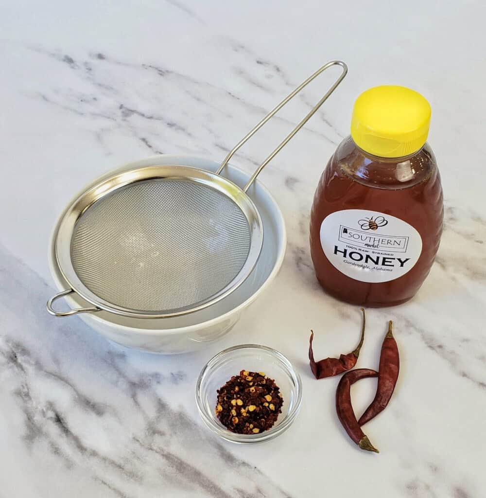 Metal sieve, bottle of honey, chile pepper flakes and peppers on marble surface