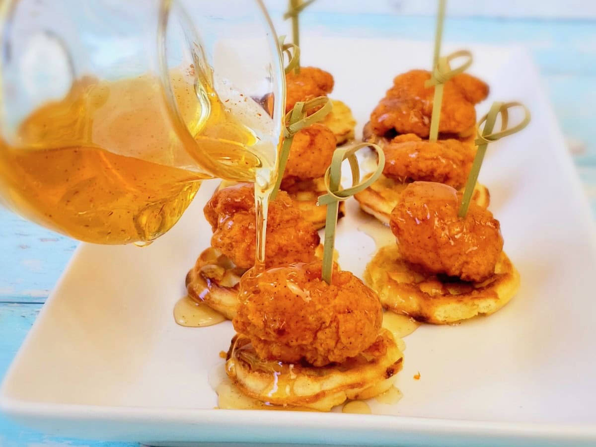 Hot honey poured from a glass pitcher onto mini waffles and chicken nuggets.
