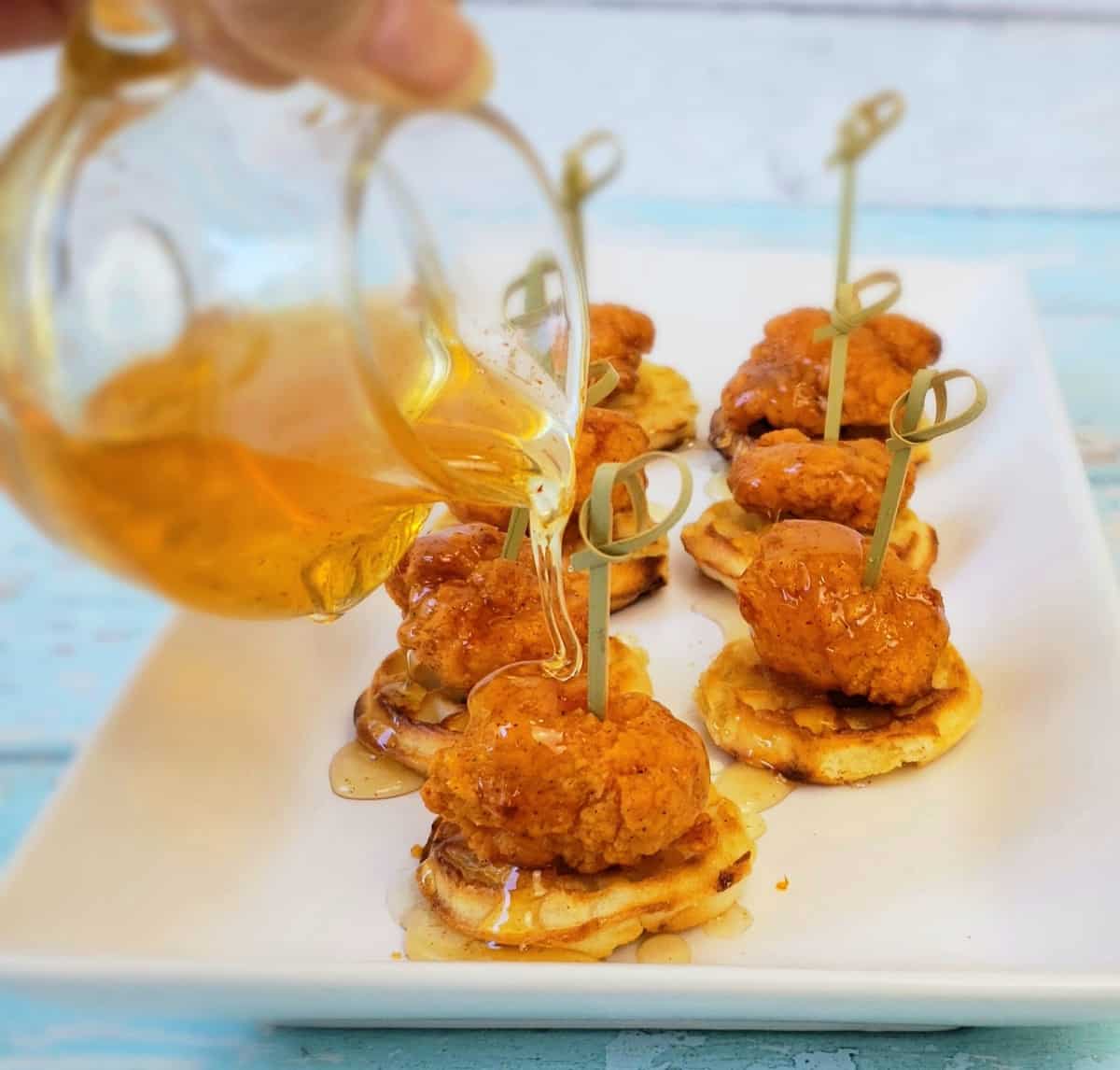 Hand pouring honey from glass pitcher onto chicken nuggets and mini waffles.