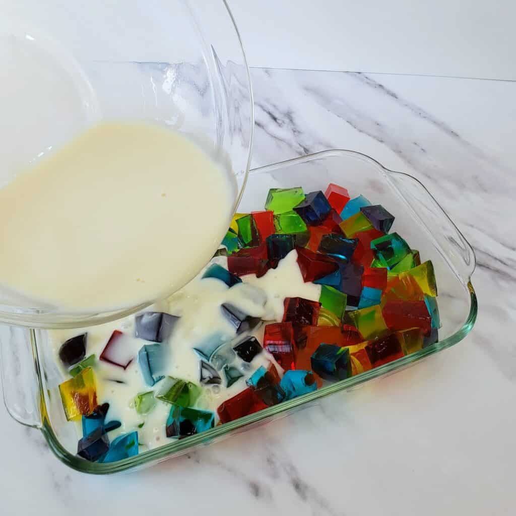 Creamy mixture being poured out of glass bowl into multicolored cubes of Jello.