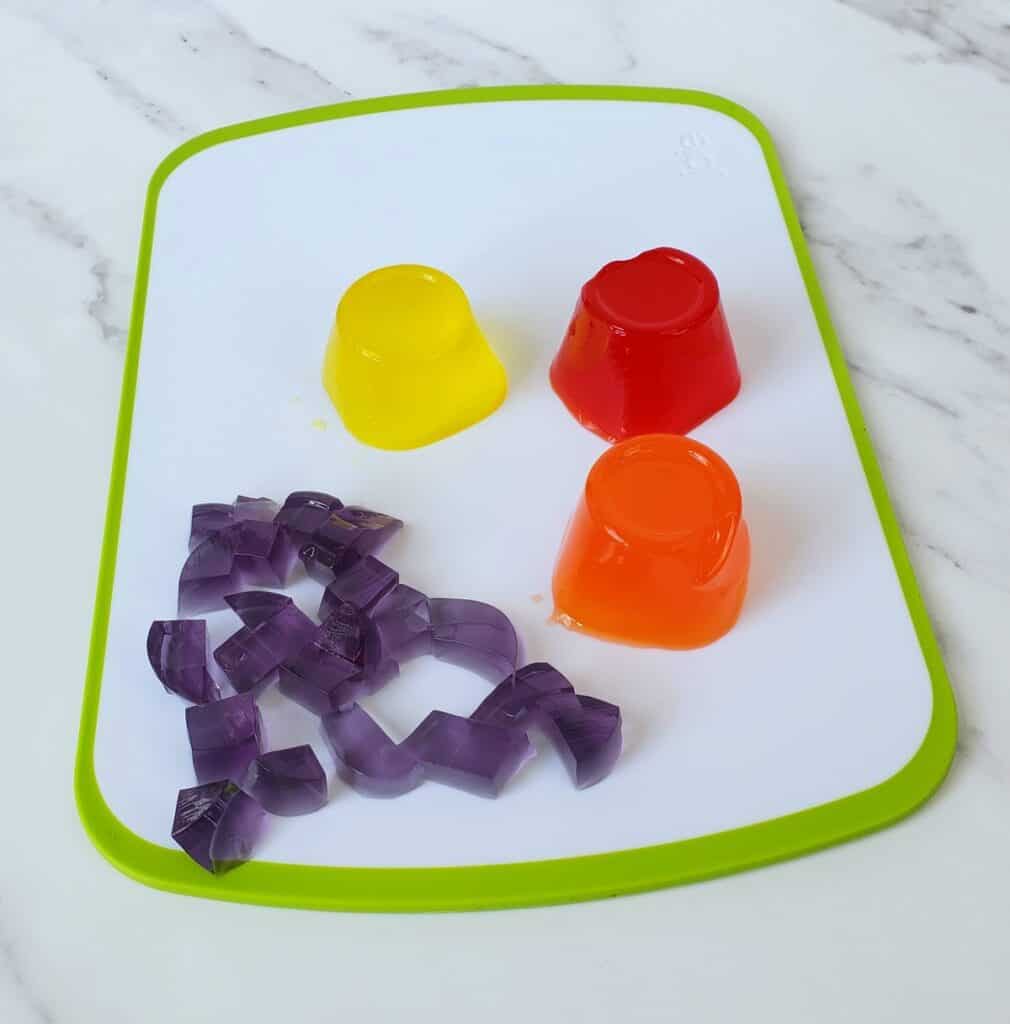 Four colors of snack pack Jello on a green and white cutting board. One of them is cubed.